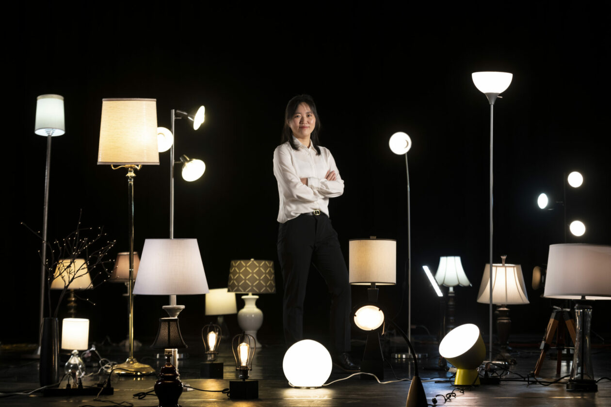 Dr. Nan Cen on a dark stage surrounded by lighted desk, table and floor lamps