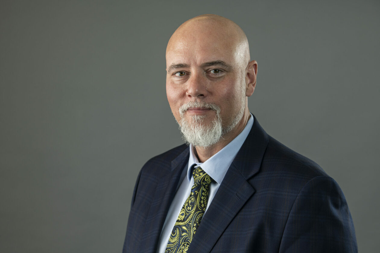 Photo of Dr. David Borrok wearing a dark suit, light blue shirt and blue and green tie. He has a shaved head and wears a goatee.