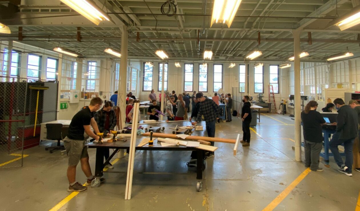 Several engineering design students gathered in a lab working with lumber and power tools in a large open area