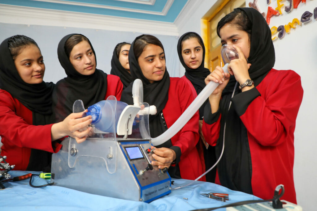 Six Afghan teenage girls wearing red jackets and black head coverings gather around a ventilator they invented.