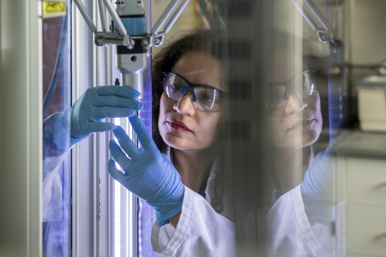 Woman wearing safety glasses and lab coat. She has long, dark, curly hair and is wearing blue latex gloves. She is looking at a long glass tube during an experiment.