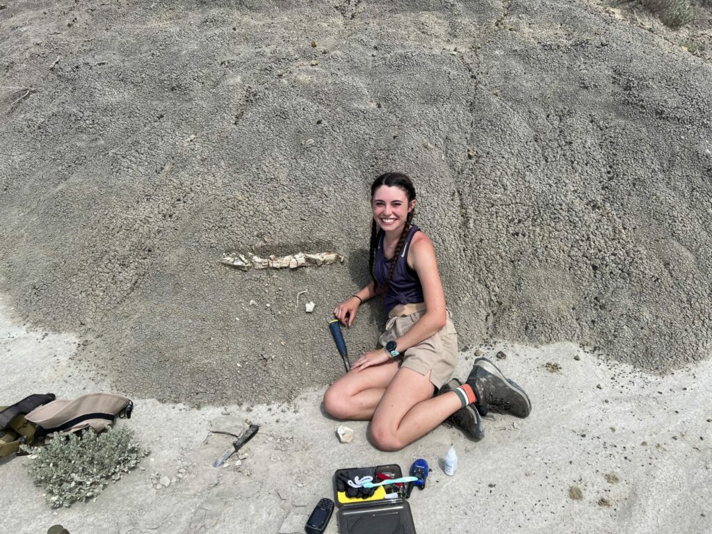 Young woman with dark hair in a braid, wearing shorts , tank top and hiking boots, sits on the ground next to the fossil she found.