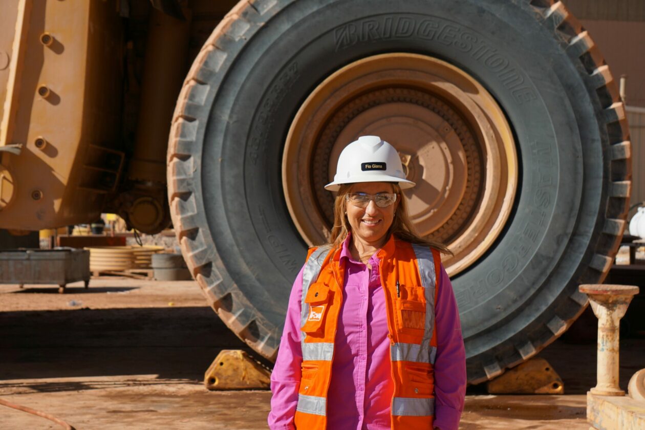 Woman in safety vest and hard hat stands beside a huge mining vehicle.