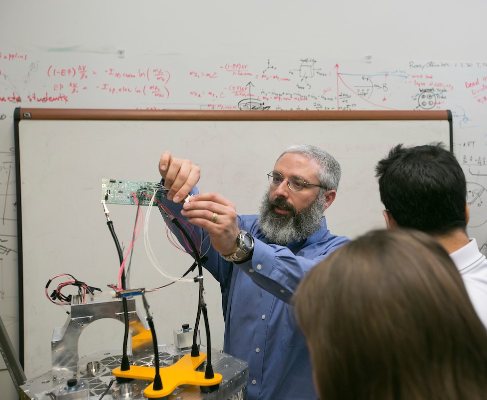 Professor and students are in a classroom working with lab equipment. The professor is wearing a blue shirt and glasses, and he has a beard.