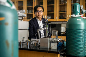 Man in dark jacket and white shirt stands in a lab surrounded by pressurized tanks and equipment