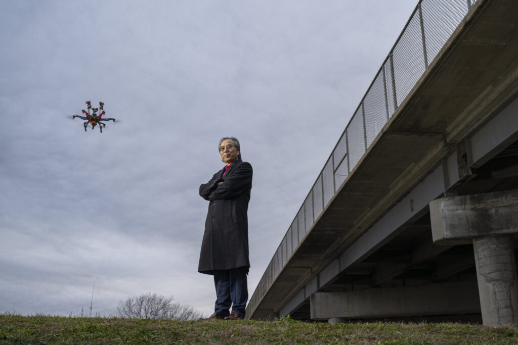 Dr. Genda Chen stands beside a Rolla, Missouri, bridge, while an unmanned aerial vehicle that is part of his BIRDS invention hovers in the sky behind him. Photo by Blaine Falkena/Missouri S&T.