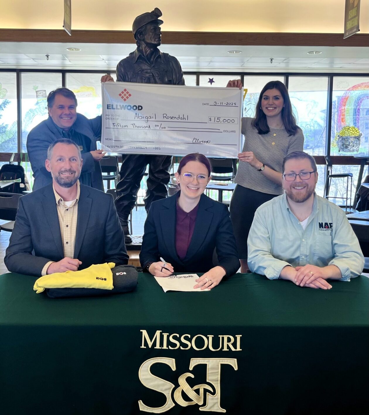 Abigal Rosendahl, front center, takes part in a ceremonial scholarship signing. Seated with Rosendahl are her father, Justin Rosendahl, left, and Calum Learn from North American Forgemasters, a division of Ellwood. Behind them, Dr. Michael S. Moats, left, S&T’s chair and professor of materials science and engineering, and Selah Saffels, the department’s office support assistant, hold an oversized check from Ellwood. Photo by Greg Edwards/Missouri S&T.