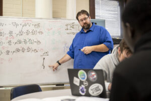 Dr. Joshua Schlegel teaches S&T students in a classroom, pointing at calculations on a whiteboard.