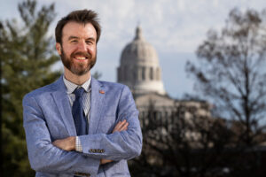 Bearded man wearing suit and tie, arms folded, stands with Missouri State Capitol building in background