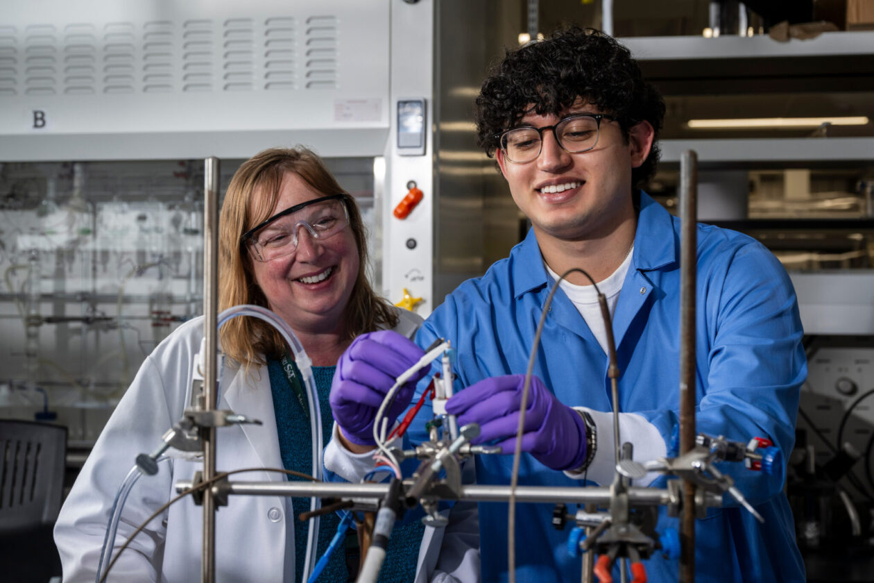 Woman scientist watches as male student uses lab equipment. Both wear goggles and lab coats.