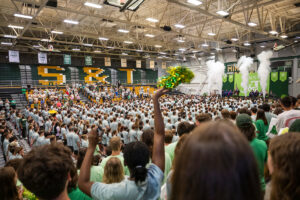 Gymnasium full of people, including new students in matching shirts. Leaders are on stage and there is a smoke/steam effect.