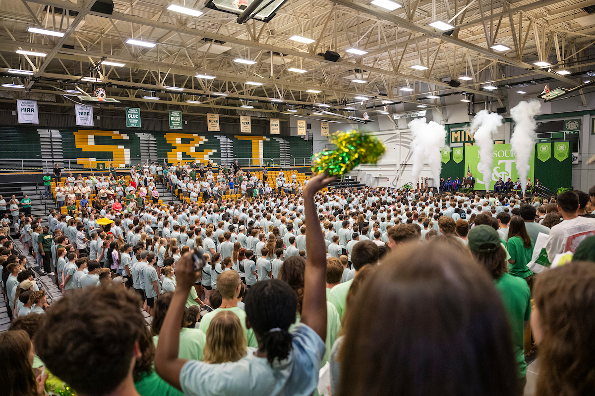Gymnasium full of people, including new students in matching shirts. Leaders are on stage and there is a smoke/steam effect.