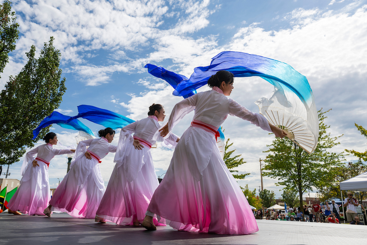 Three women in white and pink dresses, holding fans that trail blue and white fabric, performing a dance on stage at the Rolla band shell as part of Celebration of Nations in 2024.