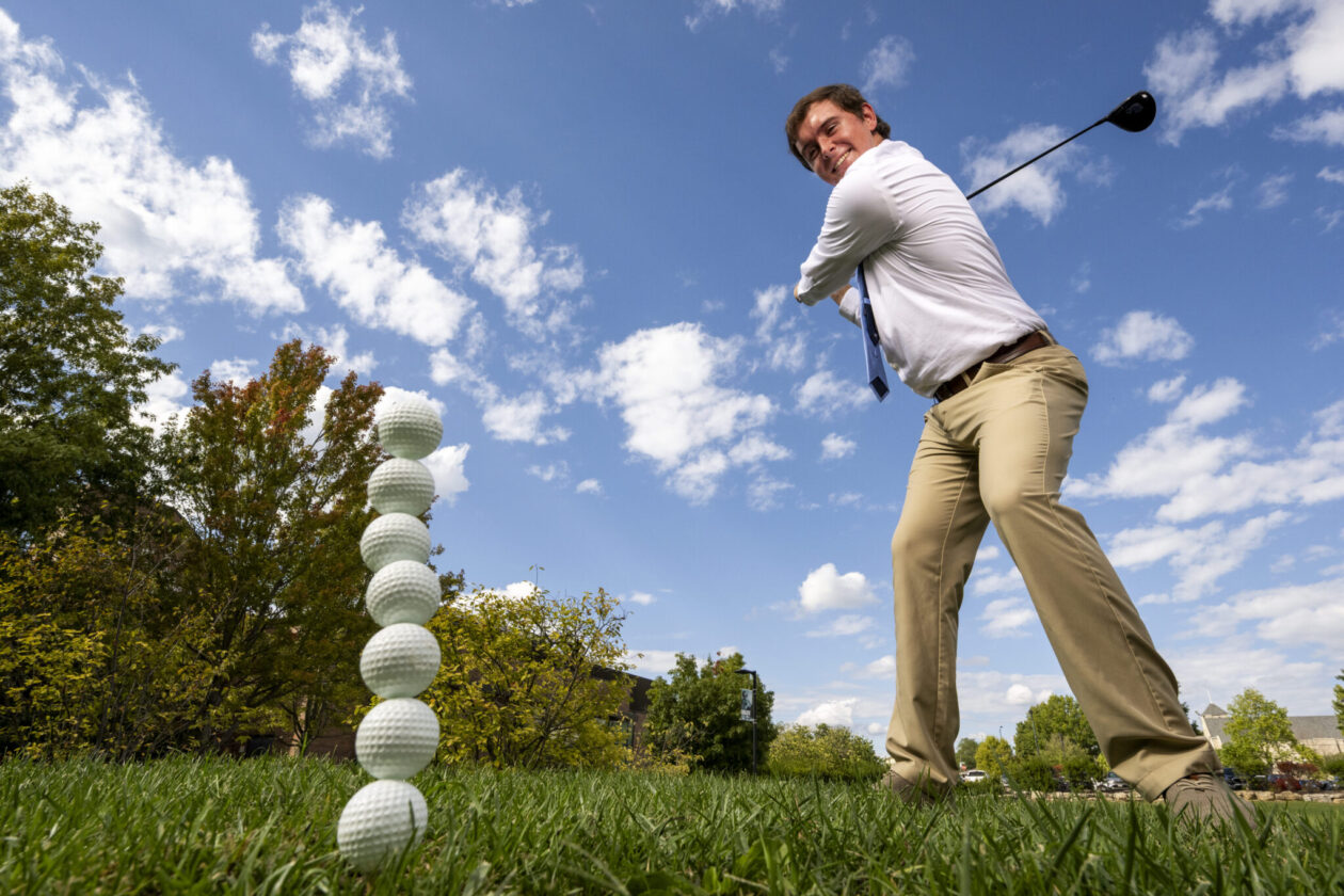 Cole Komyati swinging his golf club at a stack of golf balls on a feild.