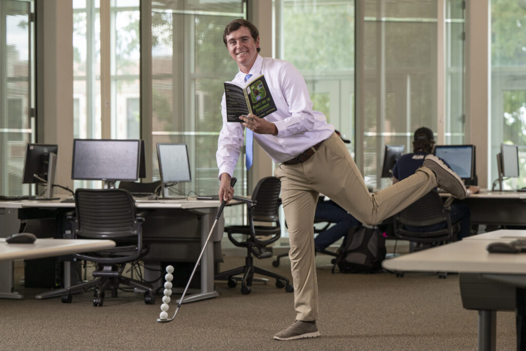 Cole Komyati balances a textbook in one hand, and a golf club with a stack of golf balls on the other in a computer lab at Missouri S&T.