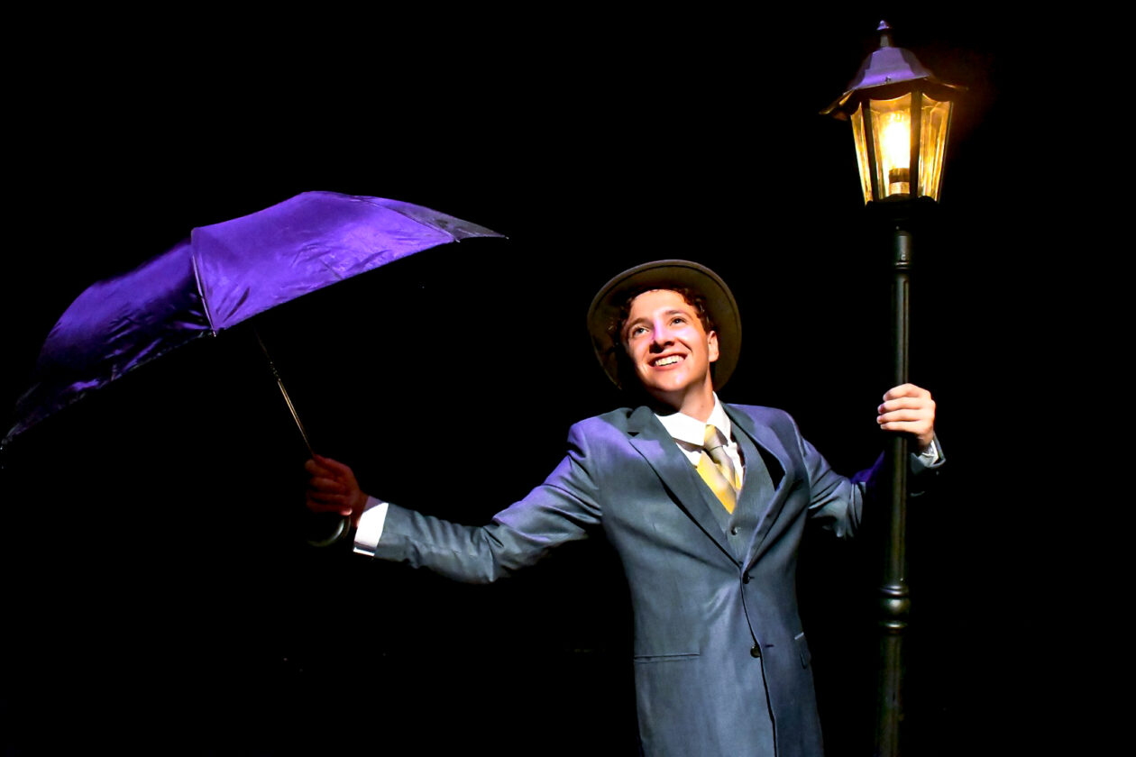 A student holding onto a lamppost and carrying a purple umbrella during a rehearsal for "Singin' in the Rain."