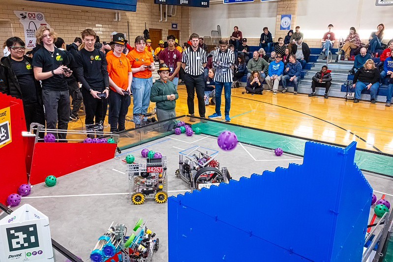 Many students gather around their robots on a platform, and two students are holding controllers; in a gym setting. In the background, a crowd watches.