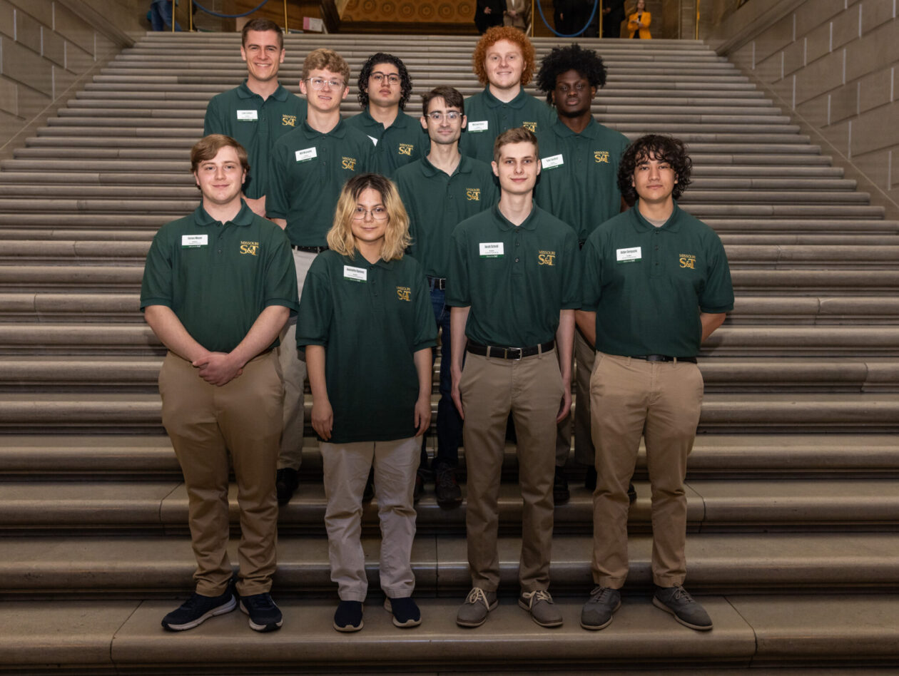 Students in green polo shirts and khaki pants stand on stairs.