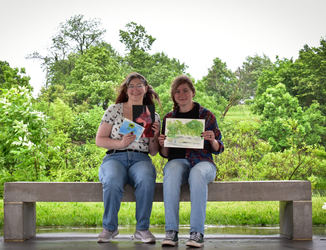 Karly Butts and Adrianna Sasser sit on a bench at a park, holding their paintings.