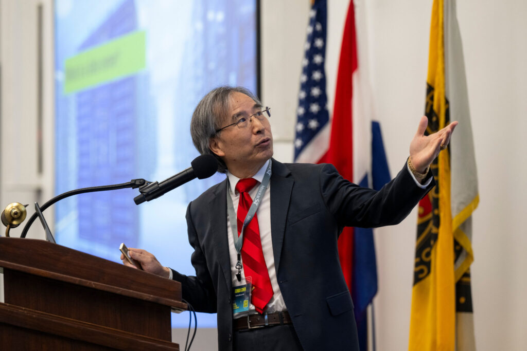 Speaker gestures to screen. He is standing at a microphone and wears a red tie. 