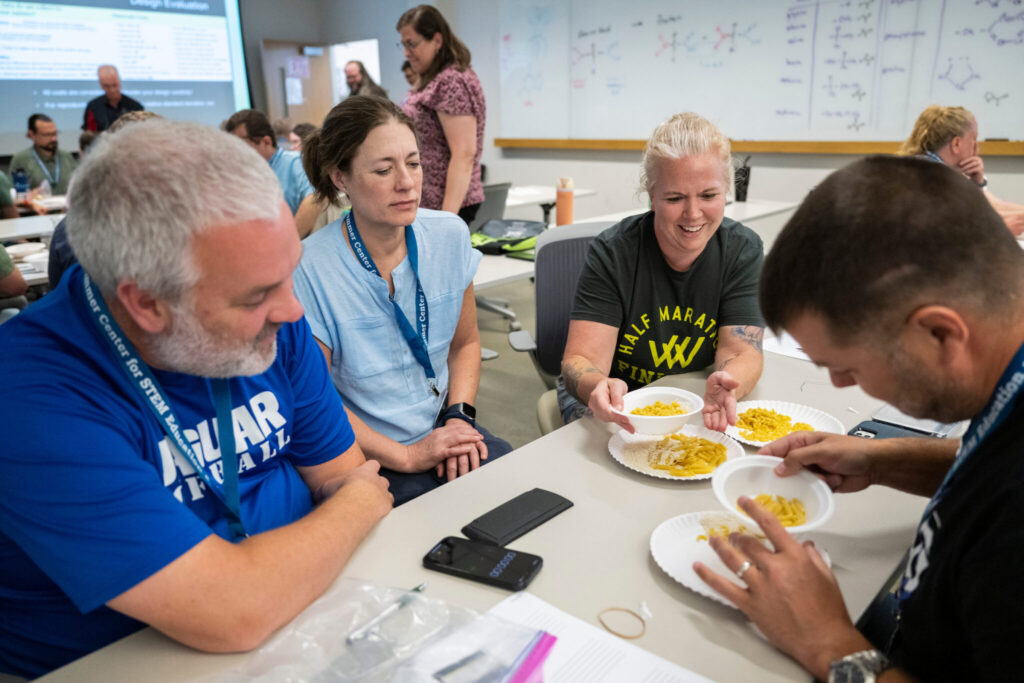 Four adults seated at a table. Two watch while two move around paper plates and pasta.