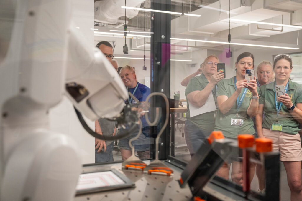 Teachers hold phones up and watch a manufacturing robot behind transparent windows.